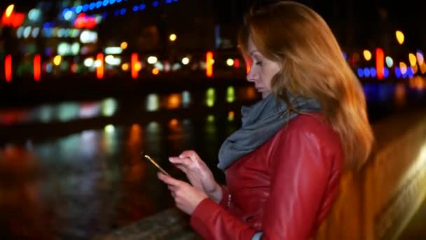 Una mujer utiliza su teléfono inteligente con una pantalla táctil. en un muelle iluminado por la noche, durante una fría noche de otoño cerca de un río, esperando a un amigo. 4k, desenfoque de fondo — Vídeo de stock