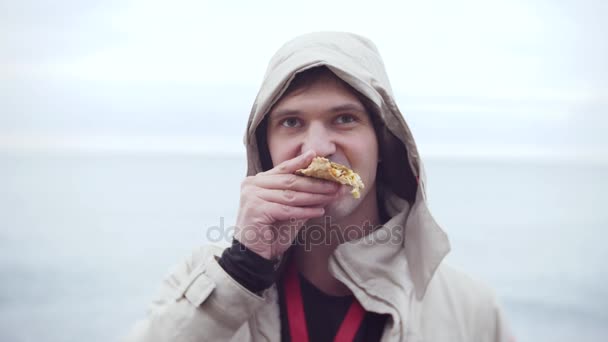 4 k. en man i en huva äter en pizza strosa längs havsstranden i kallt, molnigt väder. — Stockvideo