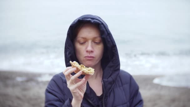 4 k. フードで女性が食べる、寒い、曇りの天気で海辺を散歩ピザ. — ストック動画