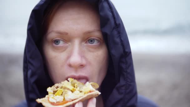 4 k. フードで女性が食べる、寒い、曇りの天気で海辺を散歩ピザ. — ストック動画