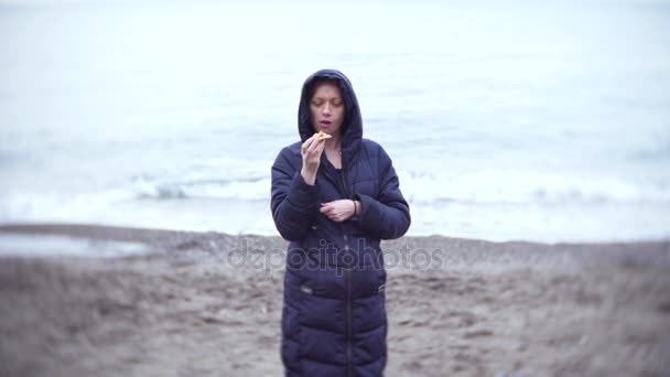 4 k. en kvinna i en huva äter en pizza strosa längs havsstranden i kallt, molnigt väder. — Stockvideo