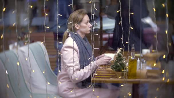 En ensam kvinna på ett café. Slow motion. Kvinnan sitter på ett kafé bakom glaset. Glas speglar stadstrafik. 4k, bakgrundsoskärpa — Stockvideo