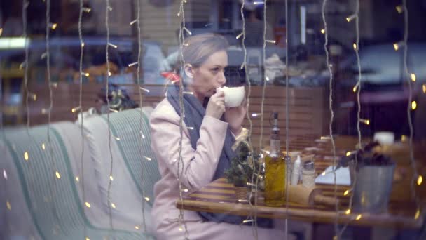 Una mujer solitaria en un café. En cámara lenta. La mujer está sentada en un café detrás del cristal. El vidrio refleja el tráfico urbano. 4k, desenfoque de fondo — Vídeos de Stock