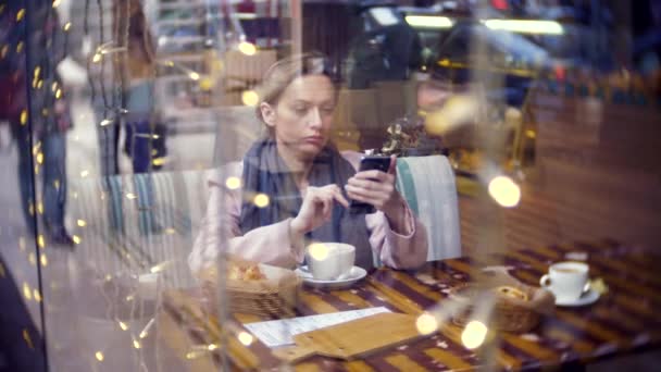 Uma mulher solitária num café. Movimento lento. A mulher está sentada num café atrás do vidro. O vidro reflete o tráfego urbano. 4k, borrão de fundo — Vídeo de Stock