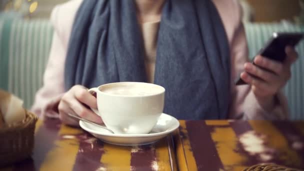 Девушка сидит в кафе с чашкой горячего кофе и зефиром. использует свой смартфон. 4k. размытие фона, боке — стоковое видео