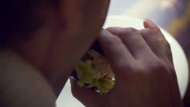 Mannen i en vit tröja som äter en svart hamburgare i en restaurang. 4k. — Stockvideo