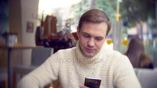 都市のカフェに座って、スマート フォンを使って白いセーターでハンサムな若い男、ウィンドウに対して。背景をぼかし。4 k — ストック動画