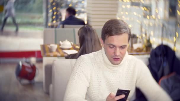 Joven guapo en un suéter blanco usando un teléfono inteligente, sentado en un café en la ciudad en un contra una ventana. fondo borroso. 4k — Vídeo de stock