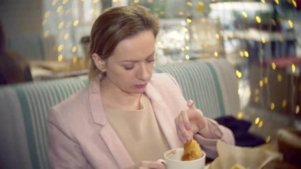 Uma menina está sentada em um café com uma xícara de café quente e marshmallow. comer um croissant. 4K. borrão de fundo, bokeh — Vídeo de Stock