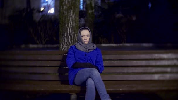 Attractive sad woman in a jacket and warm scarf sitting on a bench late at night. 4k — Stock Video