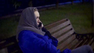 Bir ceket ve gece geç saatlerde bir smartphone kullanarak bir bankta oturan sıcak eşarp çekici üzgün kadın. Evet Shes. 4k