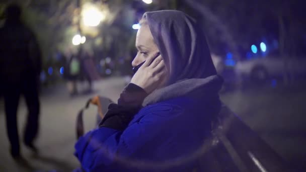 Atraktivní smutná žena v bundě a teplá šála sedí na lavičce v noci pomocí chytrého telefonu. Ona mluví po telefonu. 4k — Stock video