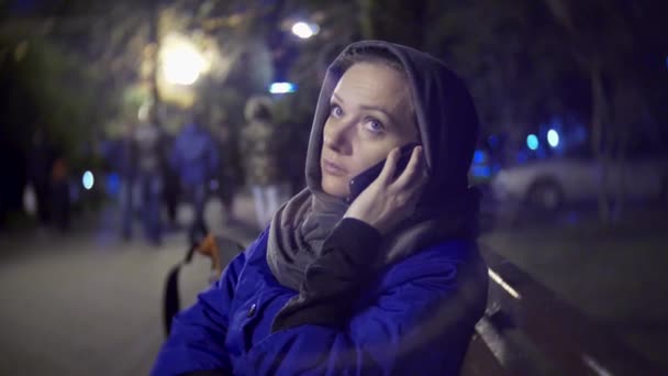 Attractive sad woman in a jacket and warm scarf sitting on a bench late at night using a smartphone. Shes talking on the phone. 4k — Stock Video