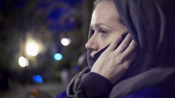 Aantrekkelijke triest vrouw in een jas en warme sjaal zittend op een bankje 's avonds laat met behulp van een smartphone. Shes praten aan de telefoon. 4k — Stockvideo