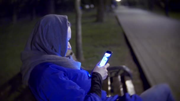 Attraktive, traurige Frau in Jacke und warmem Schal, die spätabends mit dem Smartphone auf einer Bank sitzt. Sie beschäftigt sich mit Textnachrichten, checkt E-Mails, Chatrooms oder Nachrichten im Internet. 4k — Stockvideo