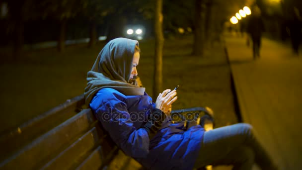 Bir ceket ve sıcak eşarp, gece geç saatlerde bir smartphone kullanarak bir bankta oturan çekici üzgün kadın. O kısa mesaj, çekleri posta, sohbet odaları veya haber Internet üzerinde çalışmaktadır. 4k — Stok video