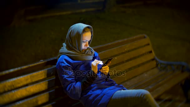 Jolie femme triste dans une veste et une écharpe chaude, assise sur un banc tard dans la nuit à l'aide d'un smartphone. Elle est engagée dans la messagerie texte, vérifie le courrier, les salles de chat ou les nouvelles sur Internet. 4k — Video