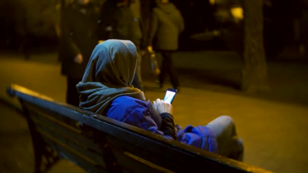 穿着夹克和暖和的围巾的迷人的悲伤的女人, 坐在长凳上深夜使用智能手机。她从事短信, 检查邮件, 聊天室或互联网上的新闻。4k — 图库视频影像