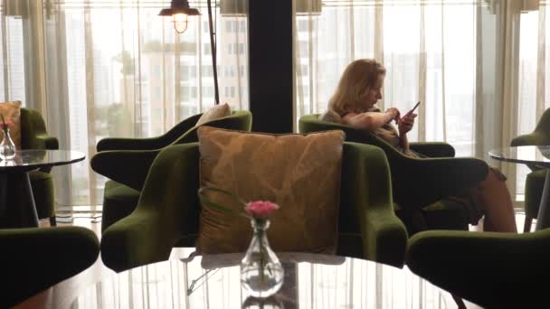金髪の女性は、高層ビルの窓からの眺め付きのカフェで携帯電話を使用します。4 k — ストック動画