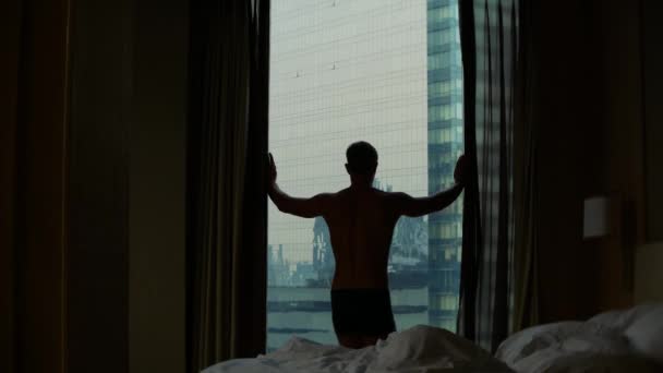 Om în pantaloni scurți deschide perdelele de pe fereastră, vedere de la fereastră la zgârie-nori. 4k — Videoclip de stoc