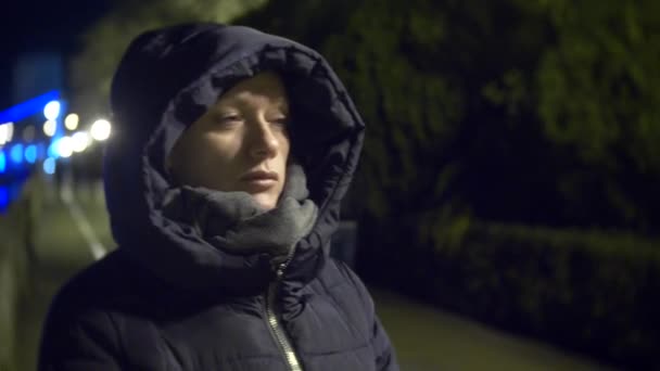 La donna è fuori la notte. Traffico urbano sullo sfondo. La ragazza con la sciarpa si congela per strada. 4k, sfocatura — Video Stock