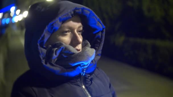 De vrouw staat buiten 's nachts. Stedelijk verkeer in de achtergrond. het meisje in de sjaal is bevriezen op straat. 4k, vervagen — Stockvideo