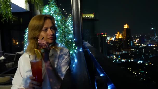 Jovem loira relaxante e bebendo coquetel no bar com vista para os arranha-céus à noite. 4k, borrão de fundo — Vídeo de Stock