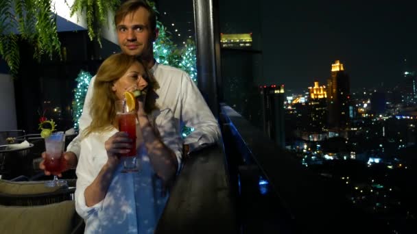 Pareja feliz abrazando, besando y bebiendo cócteles en la terraza del bar con vistas a los rascacielos, por la noche, 4k, fondo borroso. cita romántica, luna de miel . — Vídeos de Stock