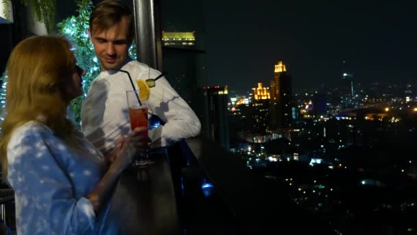 Glückliches Paar, das sich umarmt, küsst und Cocktails auf der Terrasse der Bar mit Blick auf die Wolkenkratzer trinkt, nachts, 4k, verschwommener Hintergrund. romantisches Date, Flitterwochen. — Stockvideo