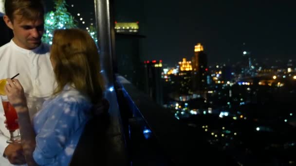 Lyckligt par kramar, Kyssar och dricka cocktails på terrassen i baren med utsikt över skyskraporna, på natten, 4k, oskärpa bakgrund. romantisk datum, smekmånad. — Stockvideo