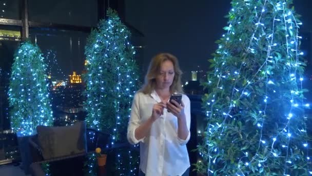 Joven y hermosa mujer rubia usando un teléfono inteligente, en una terraza del bar con vistas a los rascacielos por la noche. 4k, desenfoque de fondo . — Vídeo de stock