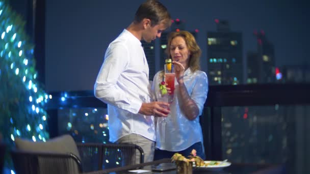 Счастливая пара обнимается, целуется и пьет коктейли на террасе бара с видом на небоскребы, ночью, 4k, размытый фон. романтическое свидание, медовый месяц . — стоковое видео