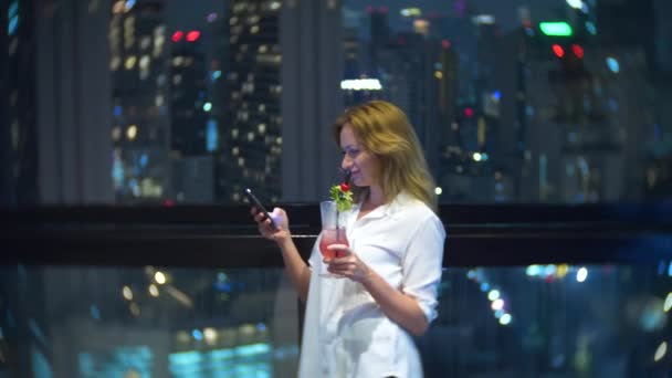 Joven y hermosa rubia usando un teléfono inteligente y bebiendo un cóctel, en una terraza del bar con vistas a los rascacielos por la noche. 4k, desenfoque de fondo . — Vídeo de stock