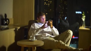 Genç, güzel bir sandalyeye gökdelenler geceleri bakan panoramik pencereli bir odada bir akıllı telefon kullanan adam. 4k, arka plan bulanıklık.