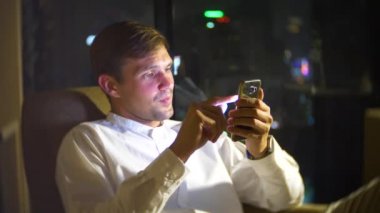 Genç, güzel bir sandalyeye gökdelenler geceleri bakan panoramik pencereli bir odada bir akıllı telefon kullanan adam. 4k, arka plan bulanıklık.
