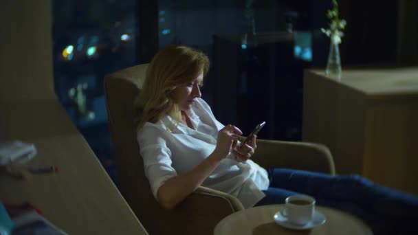 Joven, hermosa mujer rubia usando un teléfono inteligente, en una silla en una habitación con una ventana panorámica con vistas a los rascacielos por la noche. 4k, borra el fondo . — Vídeos de Stock