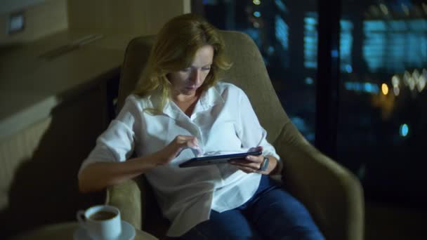Joven, hermosa mujer rubia usando un teléfono inteligente, en una silla en una habitación con una ventana panorámica con vistas a los rascacielos por la noche. 4k, borra el fondo . — Vídeo de stock