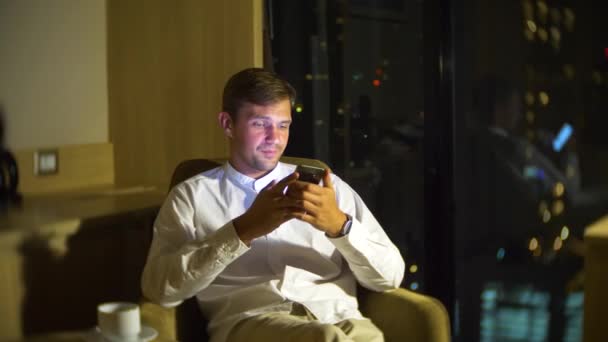 Joven y hermoso hombre usando un teléfono inteligente, en una silla en una habitación con una ventana panorámica con vistas a los rascacielos por la noche. 4k, borra el fondo . — Vídeo de stock