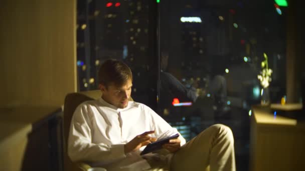 若い、美しい男、夜の高層ビルを見渡すパノラマ ビューの窓付きの部屋の椅子に、スマート フォンを使用します。4 k は、背景をぼかす. — ストック動画