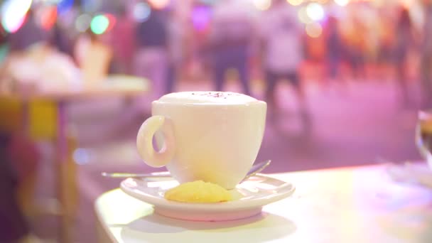 Una taza de café caliente en una mesa en un café, en una terraza con vistas a una calle peatonal ocupada. noche, contraluz, 4k, desenfoque de fondo — Vídeos de Stock