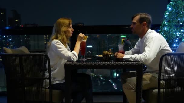 Una feliz pareja hablando y bebiendo cócteles en la terraza del bar con vistas a los rascacielos, por la noche, 4k, fondo borroso — Vídeos de Stock