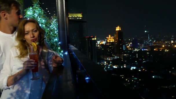 Casal feliz abraçando, beijando e bebendo coquetéis no terraço do bar com vista para os arranha-céus, à noite, 4k, fundo borrão. encontro romântico, lua-de-mel . — Vídeo de Stock