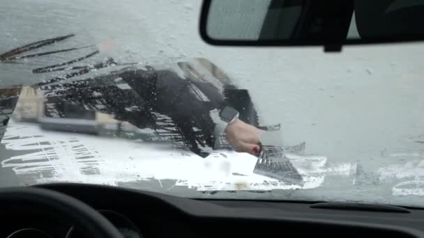 Uma mão de mulheres com uma manicura e um relógio inteligente limpa a neve da janela do carro com um raspador no inverno, uma visão do interior. 4k — Vídeo de Stock