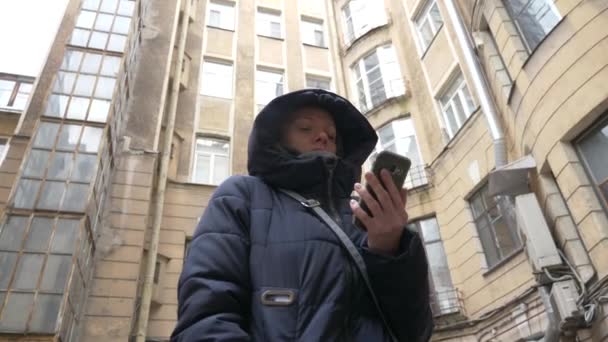 Donna che utilizza un dispositivo mobile smart phone in una giornata nevosa invernale, si trova in un vecchio cortile coperto di neve di tipo europeo. 4k . — Video Stock