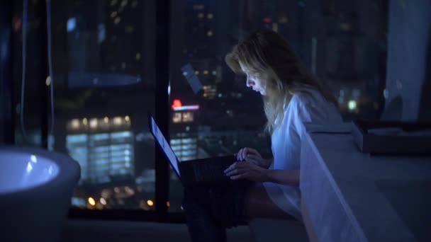 Uma mulher está sentada no banheiro em um banheiro luxuoso com uma janela panorâmica à noite com um laptop e funciona. 4k — Vídeo de Stock