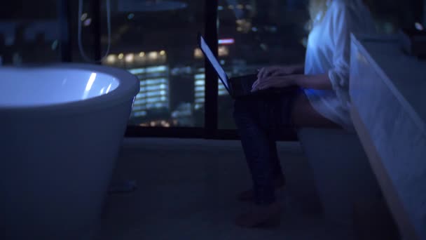 A woman is sitting in the toilet in a luxurious restroom with a panoramic window at night with a laptop and works. 4k — Stock Video