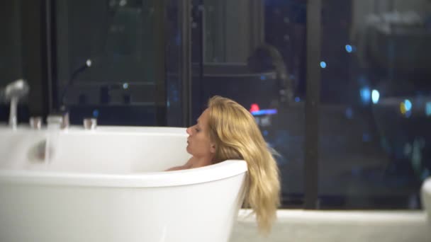 Mulher bonita desfrutando de uma banheira relaxante em um banheiro luxuoso com uma janela à noite. O conceito de um modo de vida e beleza. vista da janela para os arranha-céus. 4k — Vídeo de Stock