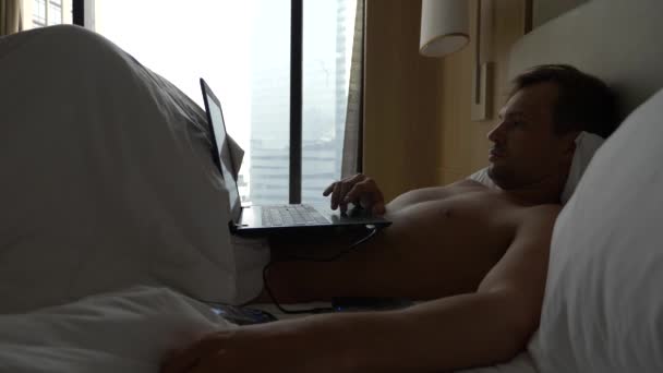 Jonge man zitten in bed met zijn open laptop en smartphone. Hes mee bezig. 4k — Stockvideo