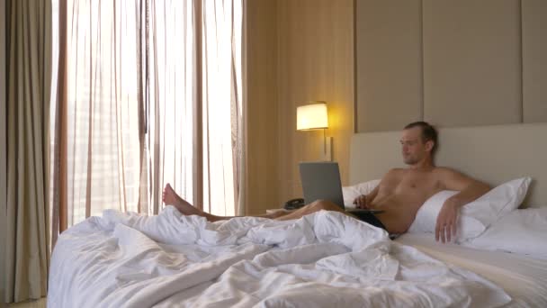 Yatakta onun dizüstü bilgisayar açık oturma ve smartphone genç adam. Hes üzerinde çalışıyor. 4k — Stok video
