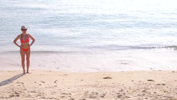 Atrakcyjna kobieta w kapeluszu stoi na plaży przed błękitne morze. Na głowie ma okulary i panama. Na jej ciało jest jasny różowy strój kąpielowy. 4k, zwolnionym tempie — Wideo stockowe